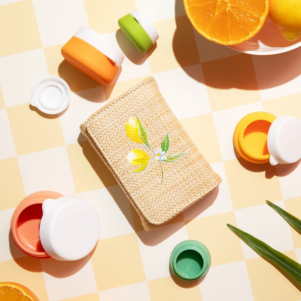 A rattan pill case with lemon print is surrounded by colorful silicone jars in orange, mango, yellow, and green. These Miamica Capri accessories are sitting on a yellow checked counter.