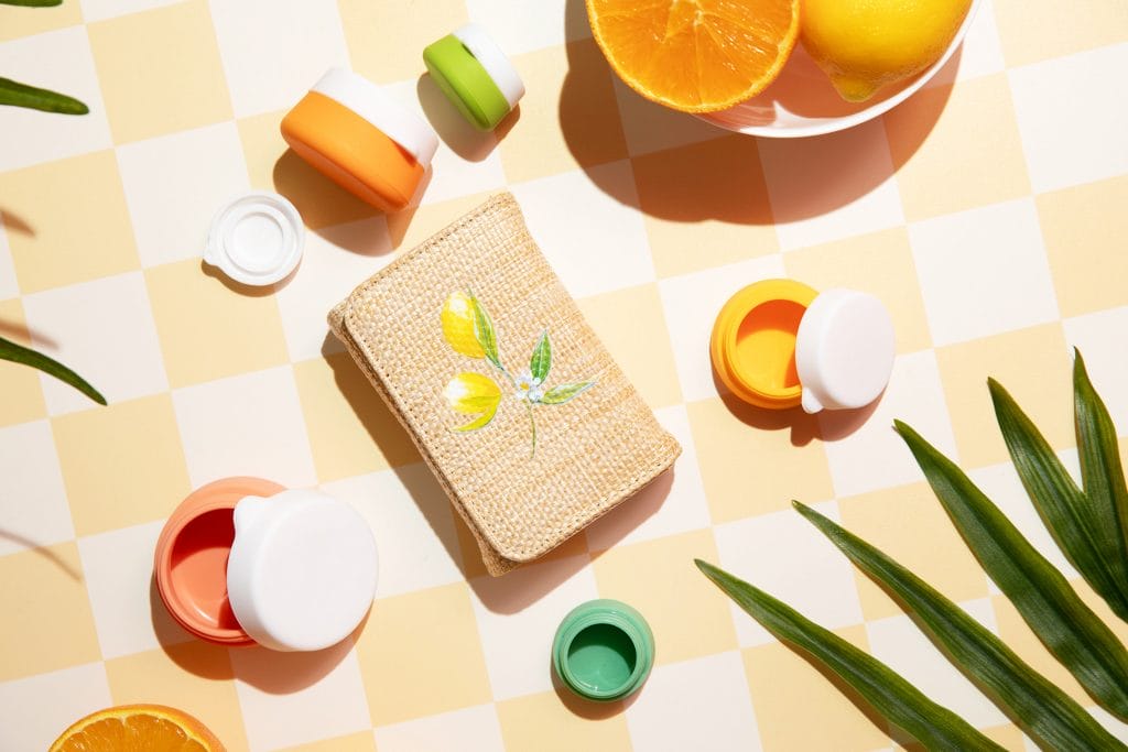 Miamica's Capri Collection travel accessories are displayed featuring a rattan pill case with vintage lemon print, and 5 citrus-colored silicone jars with white lids.
