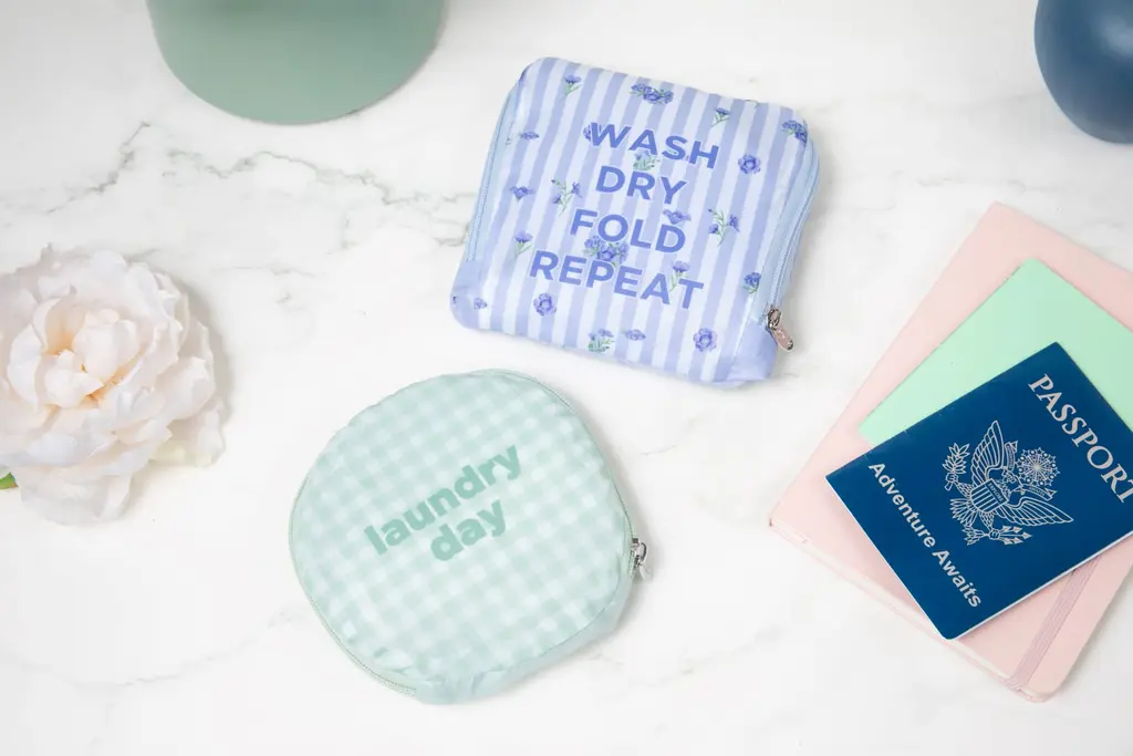Image of laundry bags from the Clubhouse Days collection. A green gingham "laundry day" bag, and a lavender "wash, dry, fold, repeat" laundry bag.